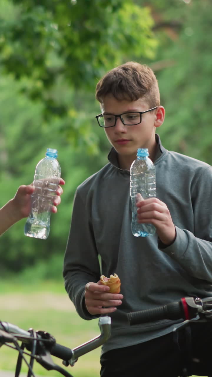 dos amigos en un paseo en bicicleta aplauden con sus botellas de agua antes de beber, sosteniendo bocadillos en sus manos, están rodeados de exuberante vegetación y árboles