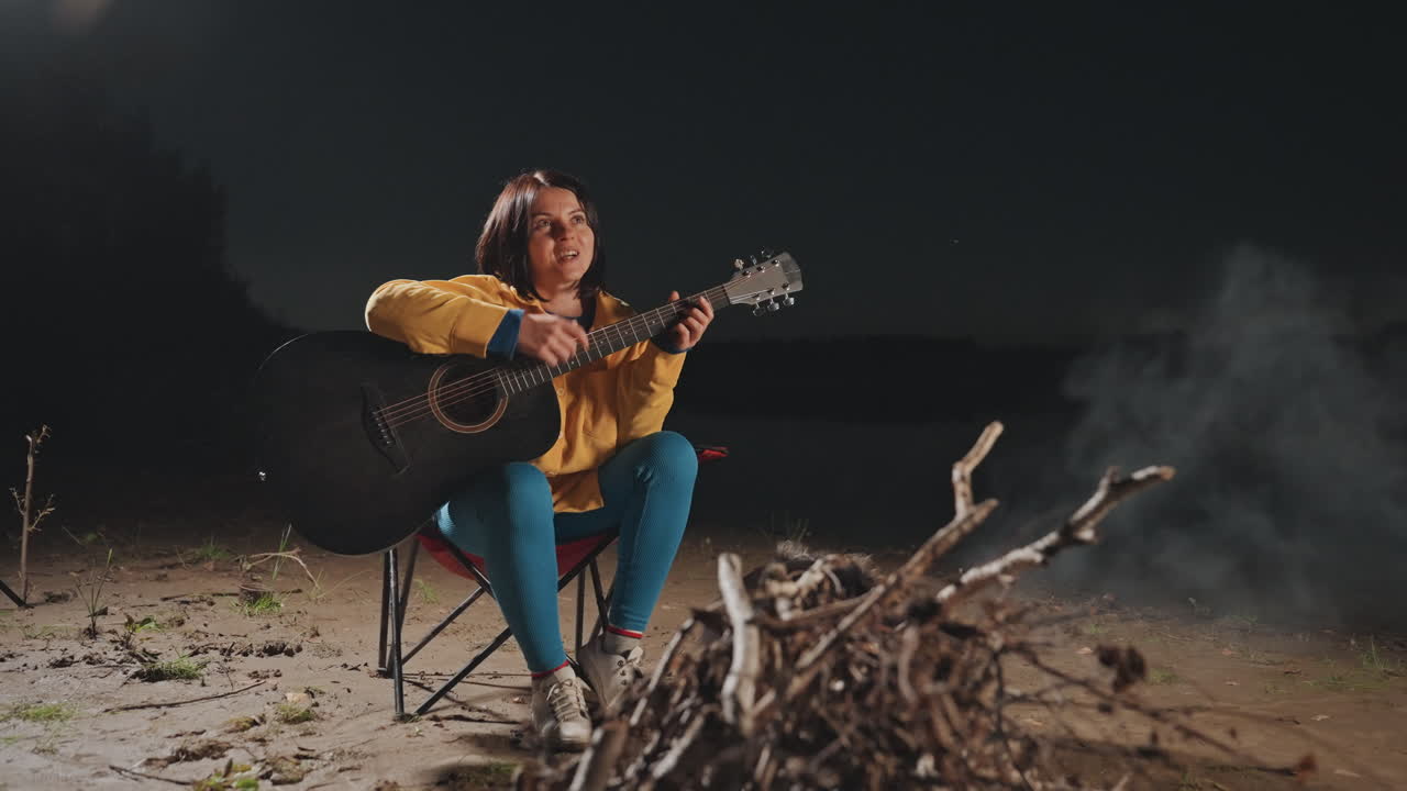 Meditativ hvid kvinde, der spiller guitar under camping, sidder ved lille bål med drivende røg og gløder, kystnat med stjernelys, introspektive vokaler og fredelig atmosfære.