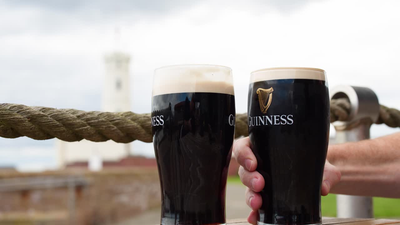Two hands clink dark beer glasses together outside, overcast daylight, shallow depth of field