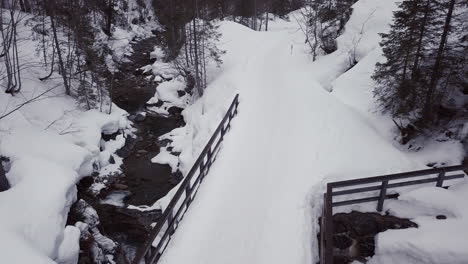 Vista-Aérea-De-Un-Pequeño-Puente-Sobre-Un-Arroyo-En-Un-Valle-Nevado-En-Los-Alpes,-Kleinwalsertal,-Austria