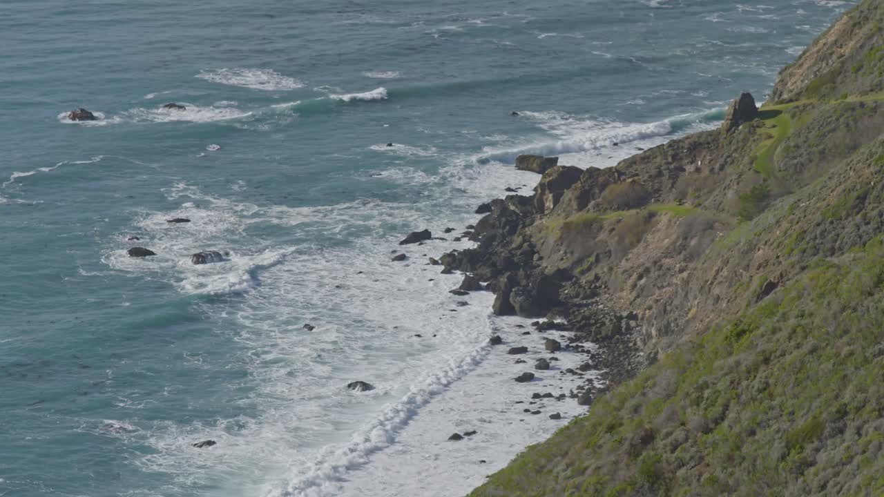 Aerial view of turquoise waves rolling against a steep, rocky shoreline along California’s iconic Big Sur coastline under soft midday light.