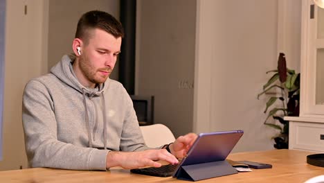 Diligent-white-young-man-enters-credit-card-information-into-tablet-computer-while-seated-at-a-table,-nonverbally-expressing-his-focus-and-attention-to-detail-with-furrowed-brows