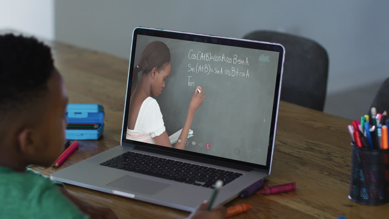 African american boy doing homework while having a video call with female teacher on laptop at home