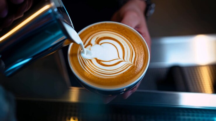 Lattiera inclinata, mani di barista che versano latte in una tazza al bancone di un caffè, formando una latte art a forma di cuore.