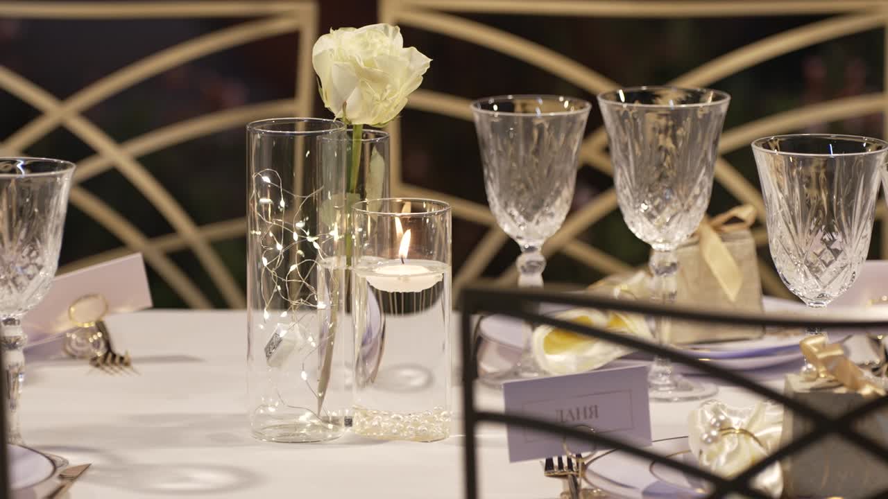 A beautifully arranged table features crystal glassware, a delicate white rose, and a glowing candle. Soft lighting creates a warm atmosphere for an evening gathering