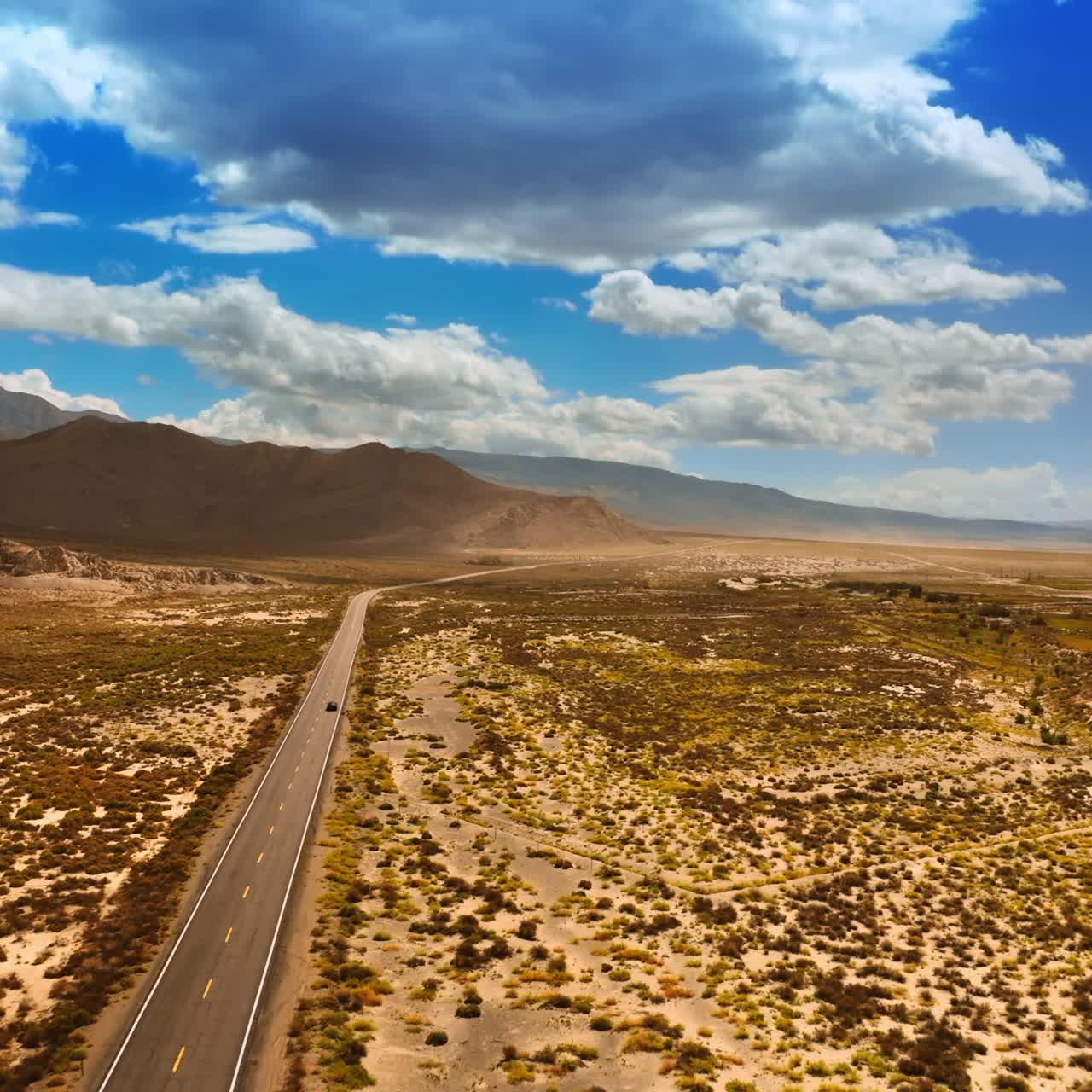 Following the lonely car riding by the highway in dry desert. Bare mountains are at backdrop. Big clouds hovering above the hot Death Valley