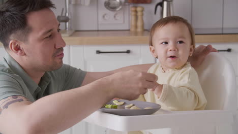 Padre-Feliz-Alimentando-A-Su-Linda-Niña-Sentada-En-Su-Silla-Alta-En-La-Cocina