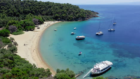 aerial: slow panning drone shot of tsougkria island beach where tourist boats and sailboats are moored while tourists swim in turquoise clear blue water