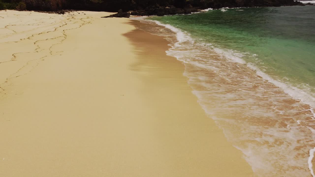 Low drone shot of soft golden sand with gentle ocean waves. Smooth upward gimbal movement reveals shoreline detail at Merese Beach near Kuta Lombok.