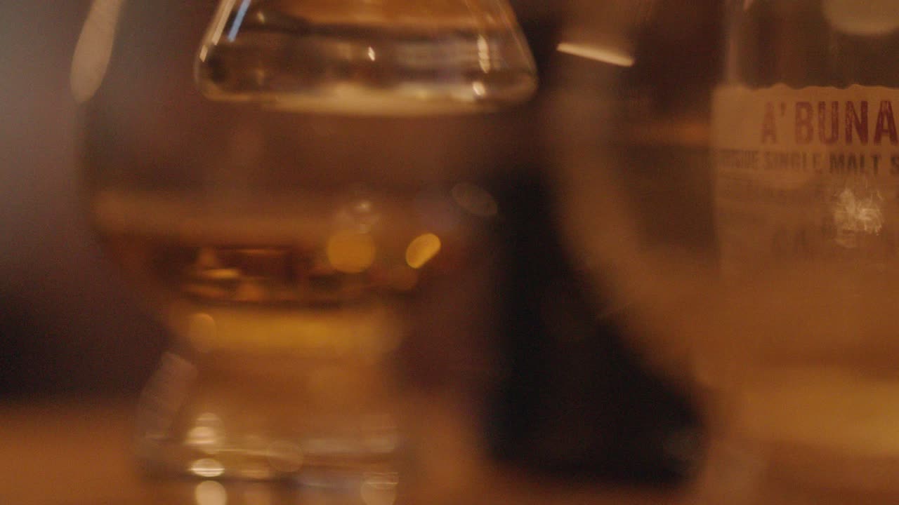 A hand reaches for a whiskey glass on a wooden bar, surrounded by other glasses in warm, ambient lighting with a shallow depth of field