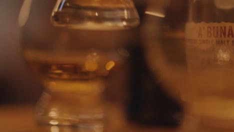 a hand reaches for a whiskey glass on a wooden bar, surrounded by other glasses in warm, ambient lighting with a shallow depth of field