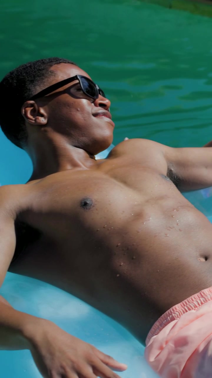 Video verticale: Un uomo illuminato dal sole su un galleggiante blu si gira verso il molo, con costume da bagno rosa e occhiali da sole scuri.