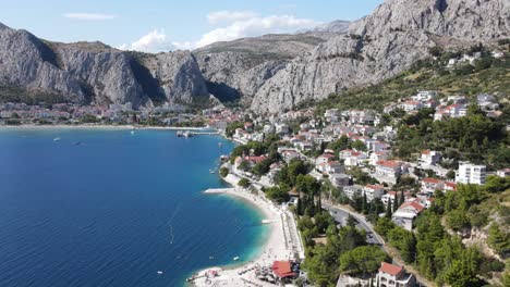 Eine-Luftaufnahme-über-üppig-Grüne-Kiefern-Und-Stahlgraue-Berge-Im-Hintergrund,-Neben-Weißem-Kiesstrand-Und-Tiefblauer-Adria,-Omis,-Kroatien