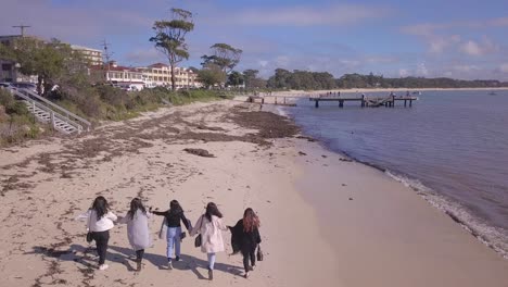 Five-young-playful-Asian-tourist-women-walk-along-the-beach,-aerial-drone-flyover-from-the-back-in-a-sunny-day