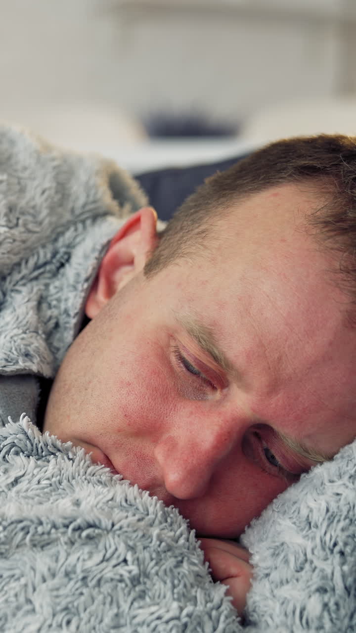Sick man with a headache during fever. Guy lying in bed covering his head with a warm blanket. Coronavirus pandemic, epidemic. Close-up. Vertical video