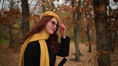 elegant lady in yellow beret and matching muffler adjusts her hair while walking through an autumn park, showcasing style and grace amidst vibrant fall colors