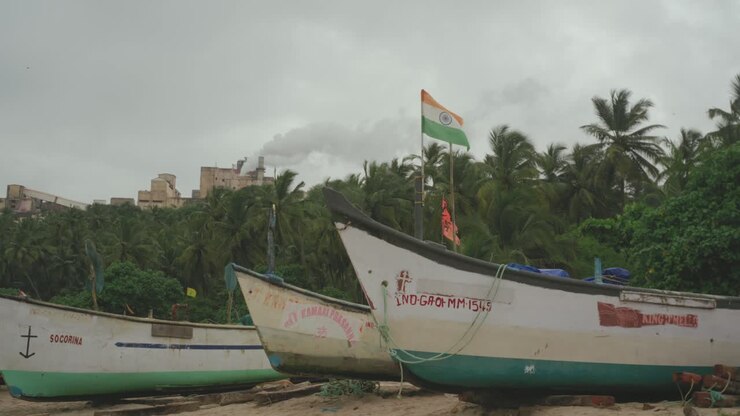 Barcos de pesca tradicionales de la India descansan en una playa tranquila bajo un cielo nublado, con una bandera india ondeando con orgullo entre las palmeras.