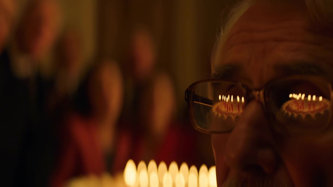A man wearing glasses is seen looking at candles in a serene and reflective indoor setting, possibly a church or formal event. The scene is captured in a close-up, slightly elevated angle.