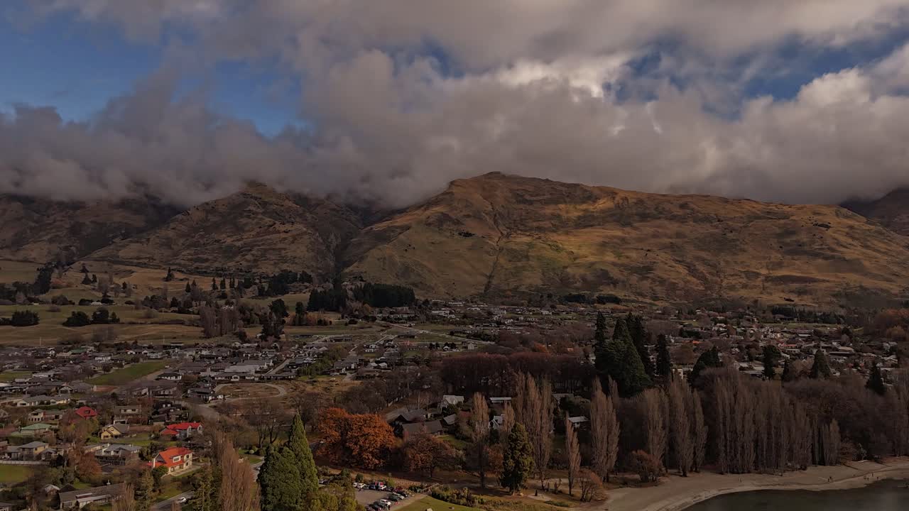 Hyperlapse drone shot over lake of wanaka town with dark clouds at sky. Colored autumn trees in fall season. Forward flight. Mountains and neighborhood in distance