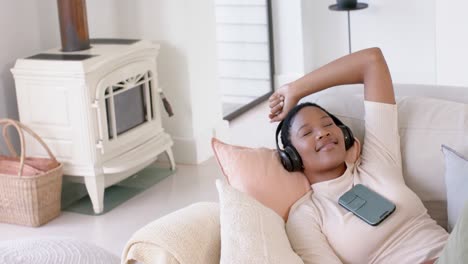 Happy-african-american-woman-lying-on-sofa-using-headphones-and-listening-to-music,-slow-motion
