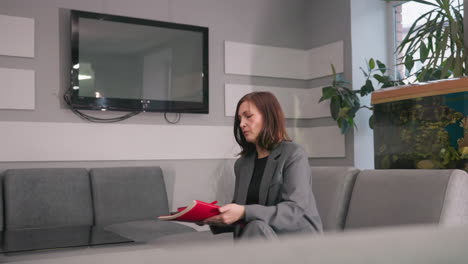 businesswoman sitting in modern office, holding red notebooks, looking down and focused. stylish workspace with plants, aquarium, and tv screen in background