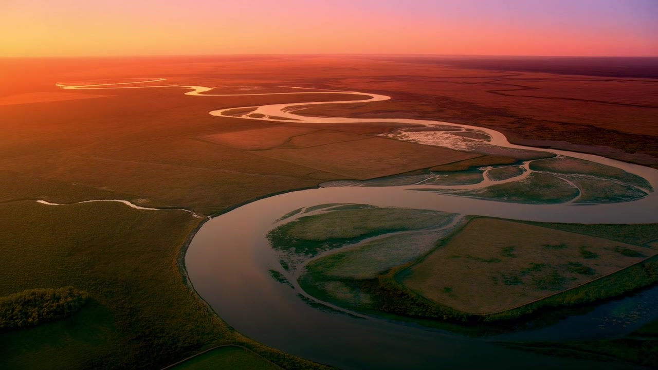 Vista aérea elevada de un río serpenteante que cruza humedales planos y campos abiertos bajo la cálida luz del atardecer con suaves reflejos y un horizonte resplandeciente.