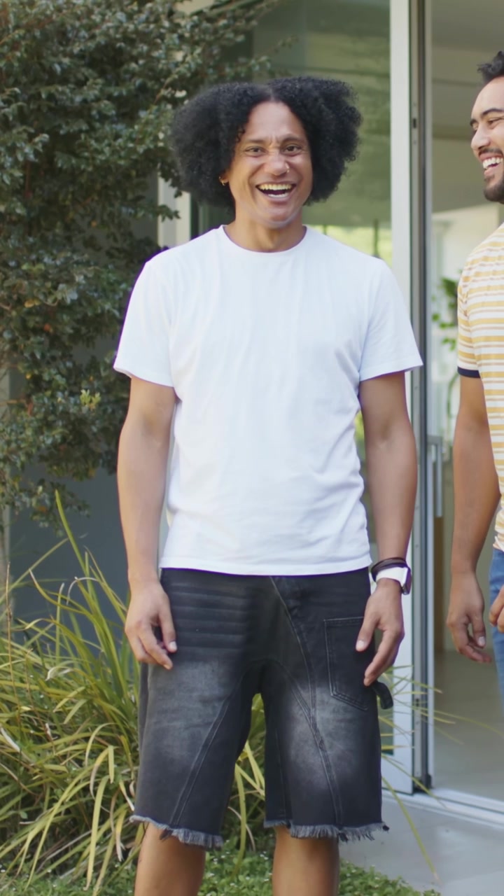 Vídeo vertical: Homem com camiseta branca saindo de casa, rindo e conversando na porta, com smartwatch visível