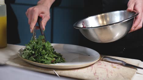 Professional-chef-building-a-salad-in-restaurant-kitchen-placing-grape-tomato-slice-from-stainless-steel-salad-mixing-bowl-onto-white-ceramic-plate-medium-tight-panning-down-slow-motion