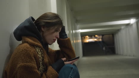 a close-up shot of a girl in a brown coat, sitting bent over in an underpass tunnel, holding her phone with a tearful expression. the scene captures an emotionally charged moment of sadness