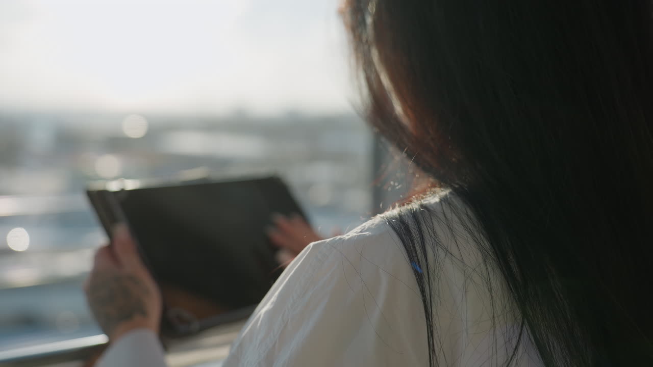 Rear view of woman with long dark hair operating tablet near window, soft sunlight creating warm glow on shoulders and glass reflecting blurred urban background with bokeh effect