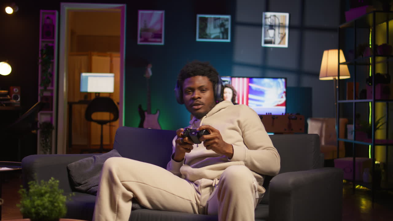 African american man playing shooter videogame in his loft apartment, holding controller for leisure activity. Male gamer participating in internet multiplayer game at night. Camera B.