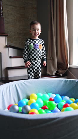 two-year baby boy pulls the dry pool with balls by the room. kid raises on the stair and drops himself into the basin. vertical video.