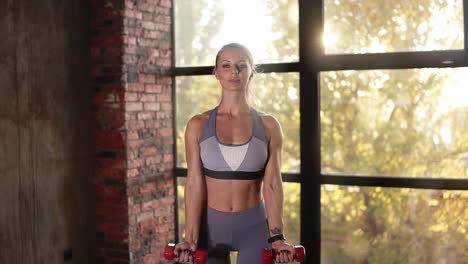 Female-athlete-or-trainer-training-with-small-red-dumbbells-in-empty-fitness-club.-Young-woman-trains-hands-using-weight-in