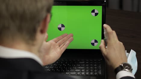 hombre se quita la máscara médica, toma desinfectante y usa cerca de la computadora portátil con pantalla verde