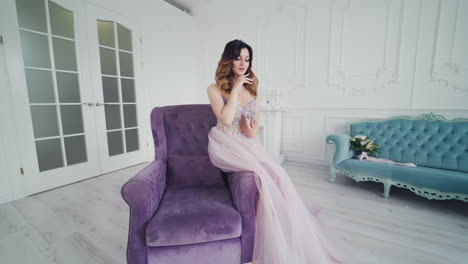 Beautiful woman model in a wedding dress sit on a chair in the spacious room. Young attractive lady in a beautiful evening dress of lavender color with pebbles color sits armchair.
