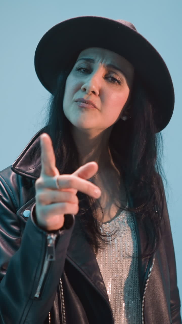 A woman in a black leather jacket, trousers, and hat sings passionately, moving her body, hands, and head. The camera zooms in slowly, against a blue background