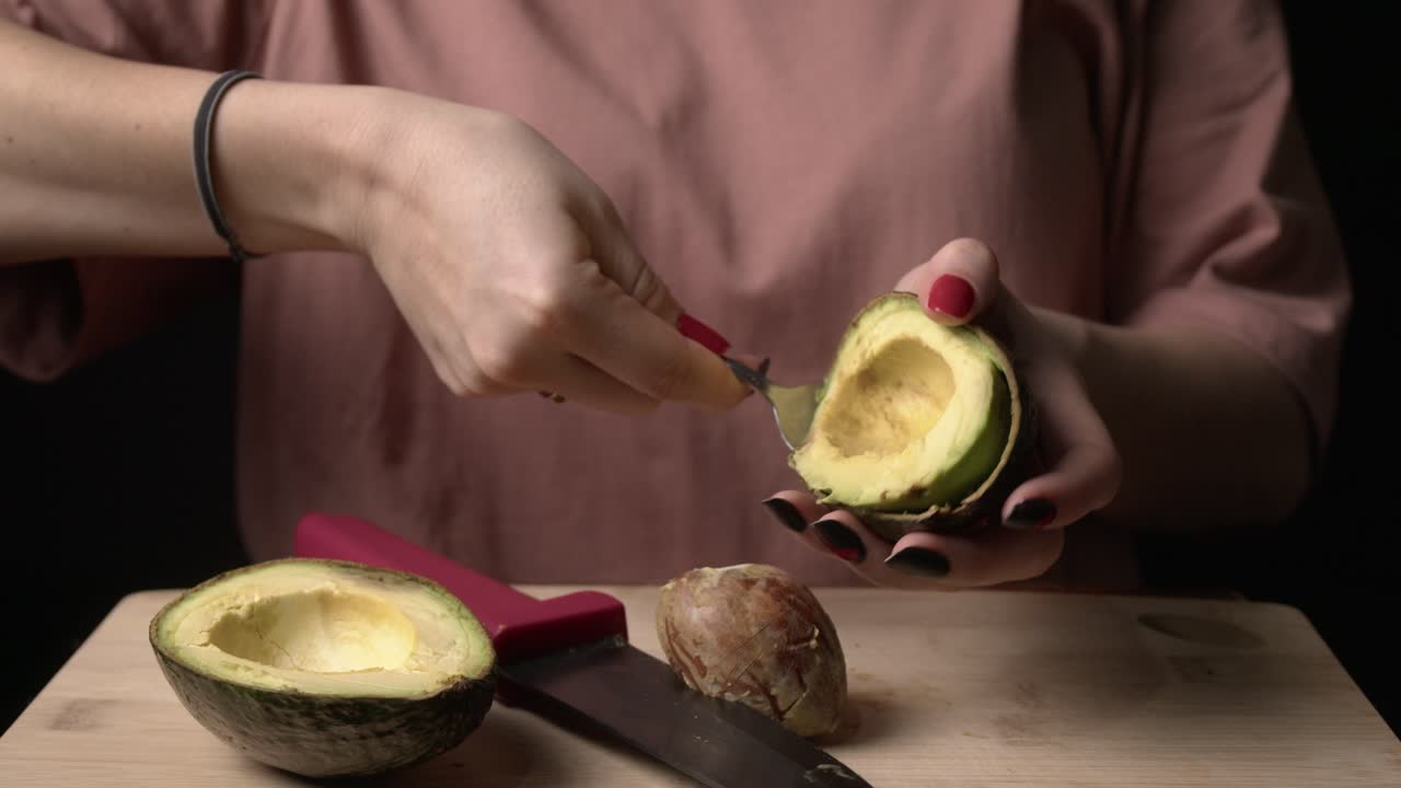 Primo piano di una donna che estrae la polpa matura di avocado dalla buccia con un cucchiaio, posato su una superficie di legno in una cucina poco illuminata.
