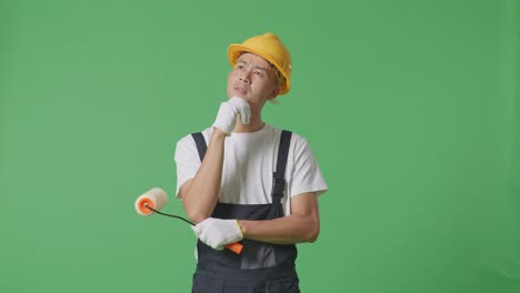 Asian Man Painter Wearing Safety Helmet Thinking About Something And Looking Around While Standing In The Green Screen Background Studio