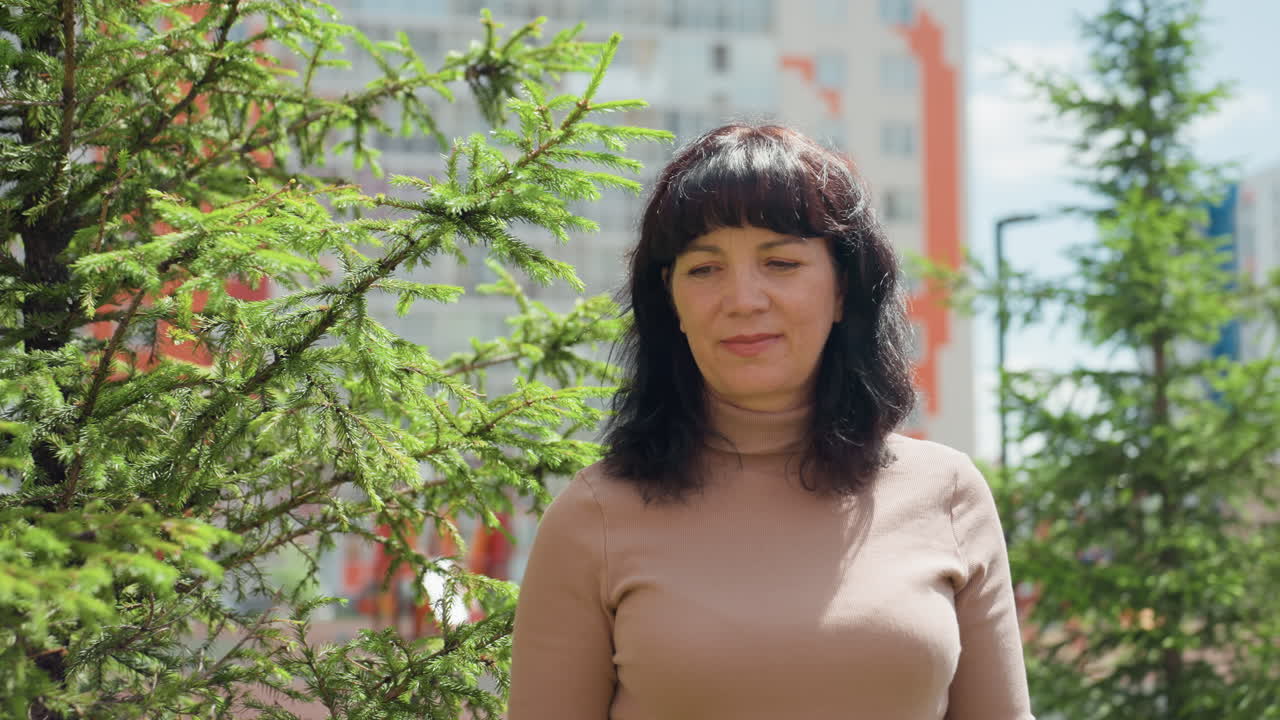 Caucasian Woman Portrait In Urban Garden, Soft Sunlight On Spruce Branches, Apartment Blocks In Background, Gentle Smile, Relaxed Posture, Gardening Hobbyist Energy, Spring Warmth And Peaceful Mood