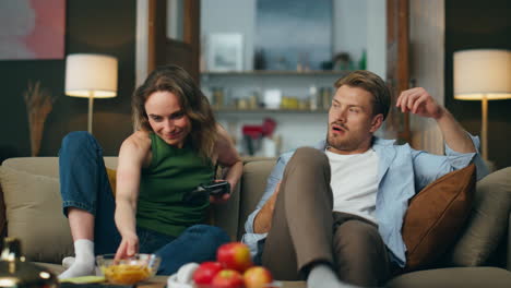 smiling woman giving snacks to man at sofa. loosing guy taking remote controller