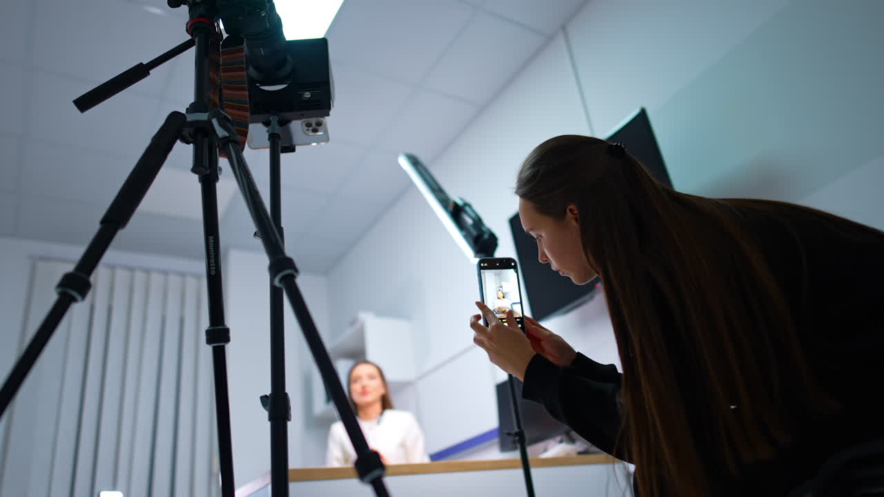 A blogger bent to catch low angle view of the speaker sitting at desk. Taking video for a blog content. Blurred backdrop.