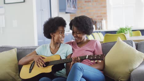 Happy-african-american-mother-and-daughter-sitting-on-sofa-playing-guitar