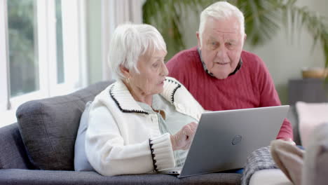 una feliz pareja caucásica sentada en el sofá usando una computadora portátil en casa, en cámara lenta