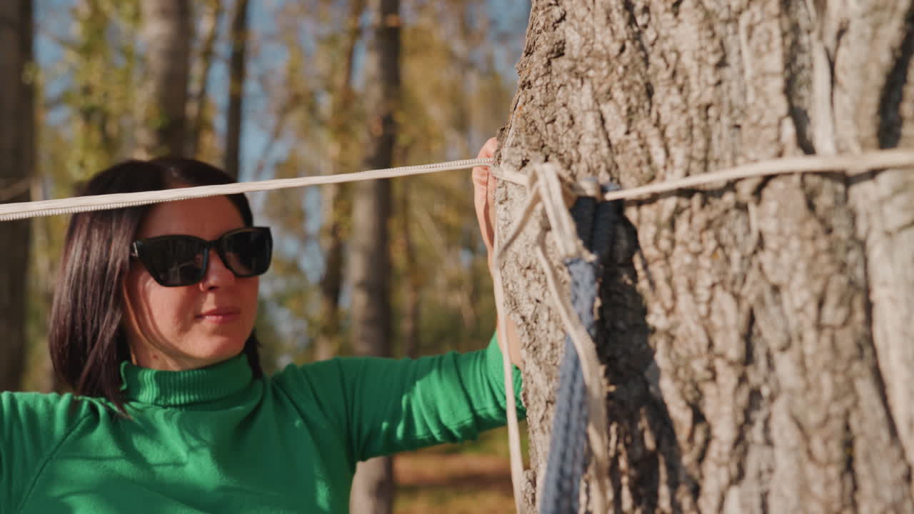 Vrouw knoopt hangmatkoord rond boom als reisblogger en weekendkampeerder, ze knoopt touw en test spanning, draagt groene trui en zonnebril, close-up handen op ruwe bast in herfstbos.