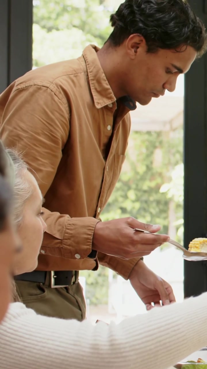 Vertical video: Serving male host in tan shirt leaning forward, spooning salad onto plates at home