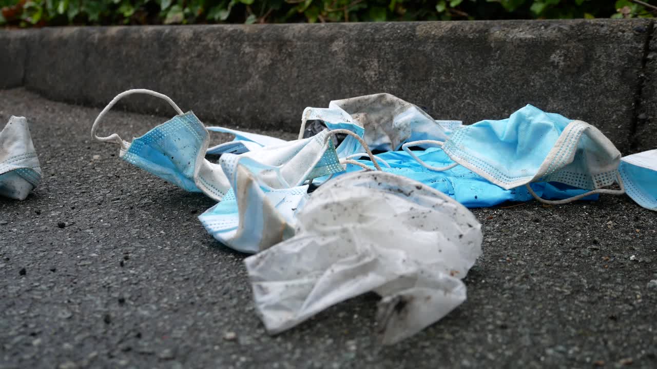 거리 보도에서 바람에 쓰레기를 불고 있는 더럽고 버려진 코로나 바이러스 마스크는 돌리가 느리게 왼쪽으로 닫힙니다.