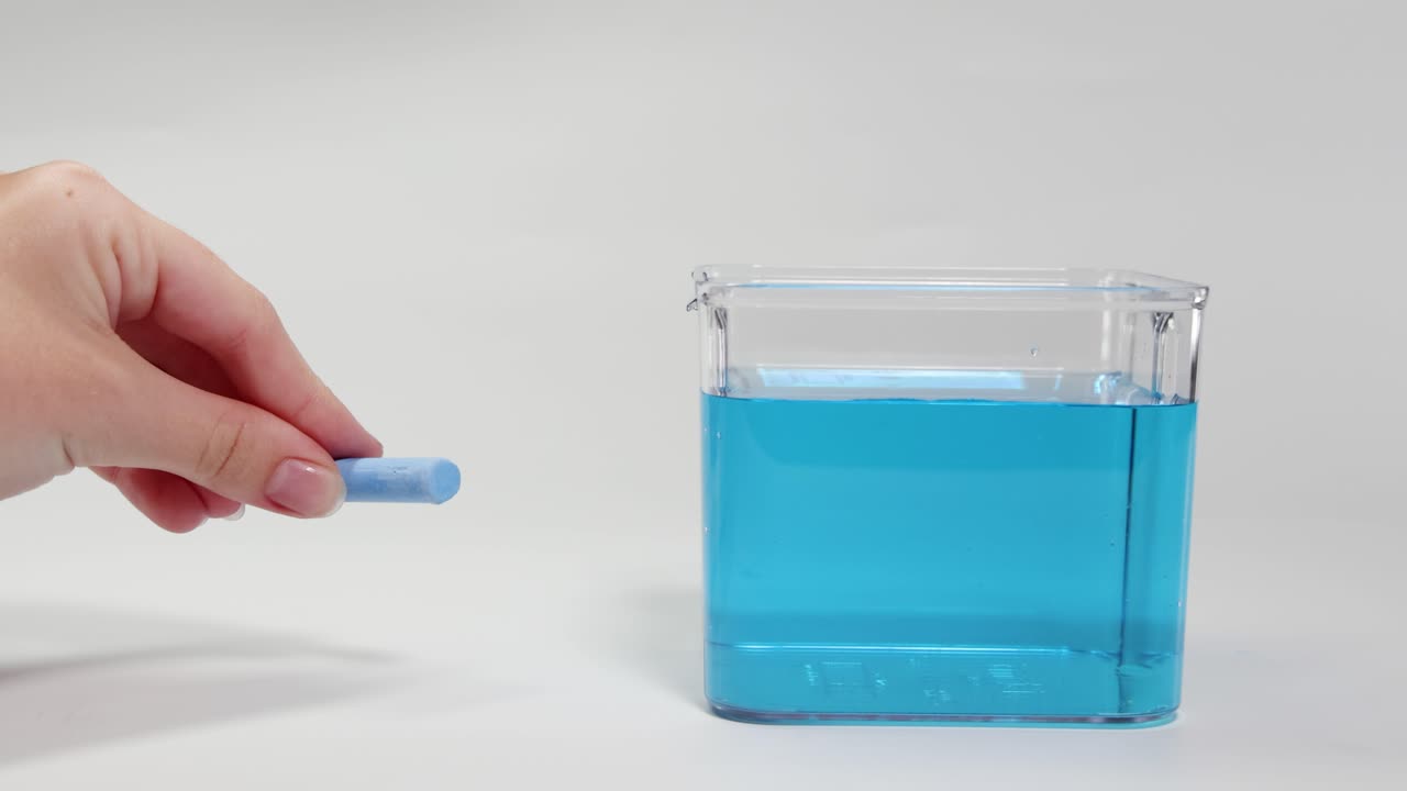 A hand drops chalk into a water-filled container, observing its sinking behavior under controlled lighting