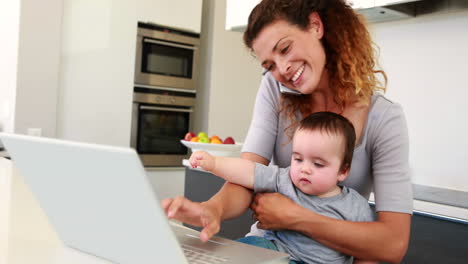 madre sentada con el bebé en el regazo usando la computadora portátil y hablando por teléfono