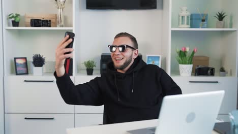 A-young-man-sitting-at-a-desk-in-the-office-with-a-laptop,-taking-a-selfie-on-his-phone-while-wearing-sunglasses-and-flashing-a-two-finger-gesture