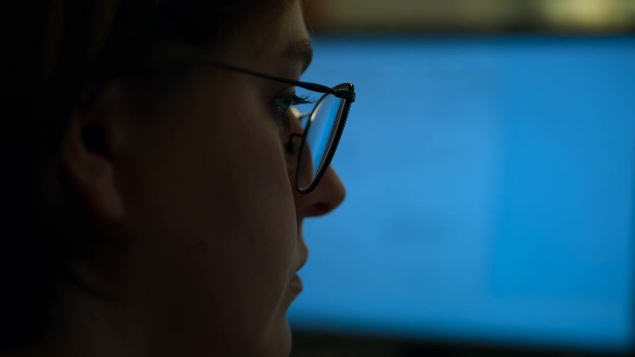 Eye-level close-up shows person in profile wearing round eyeglasses while turning head toward bright blue monitor in dim workspace, with focused gaze, soft rim light, and calm concentration